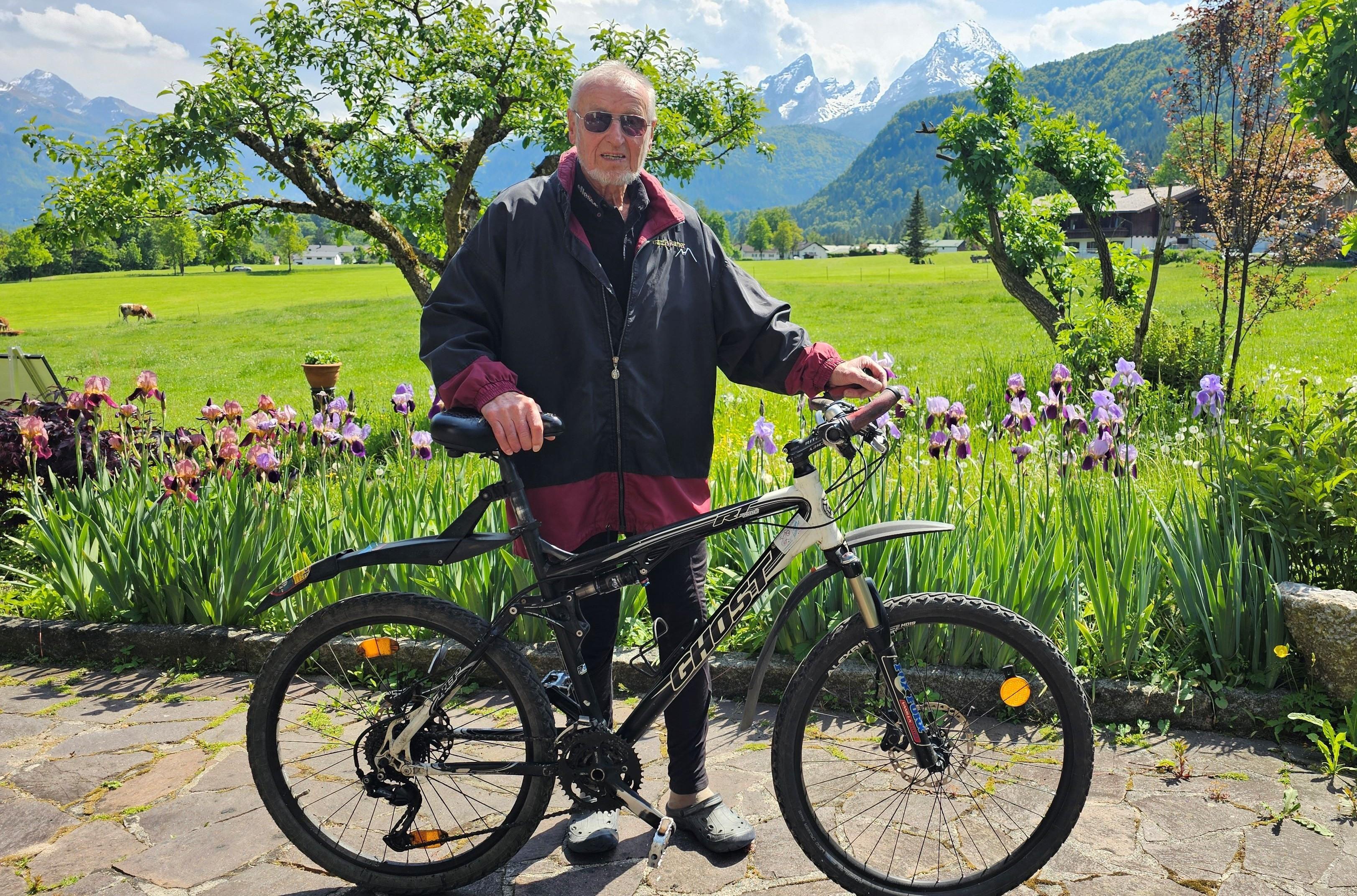 Ein alter Mann mit einer dunklen Jacke steht mit seinem Fahrrad in einem Garten, im Hintergrund sind schneebedeckte Berge zu sehen.